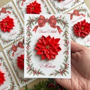 Könnte beinhalten: Eine rechteckige weiße Karte mit einer roten Weihnachtssternblüte, einer roten Schleife und einem grünen Kranz aus Stechpalmen. Der Text Buon Natale und der Name Marco sind in Schreibschrift gedruckt, inklusive des Logos Valentina.