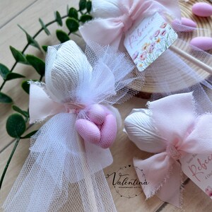 K&ouml;nnte beinhalten: Nahaufnahme von handgefertigten Gastgeschenken mit wei&szlig;em T&uuml;ll, rosa Schleifen und dekorativen Elementen. Jedes Gastgeschenk enth&auml;lt ein wei&szlig;es, strukturiertes tulpenf&ouml;rmiges Element und rosa S&uuml;&szlig;igkeiten. Die Gastgeschenke sind mit kleinen Etiketten mit floralen Designs und dem Namen "Rebecca" verziert.