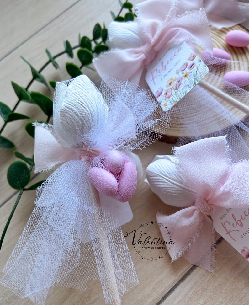 K&ouml;nnte beinhalten: Nahaufnahme von handgefertigten Gastgeschenken mit wei&szlig;em T&uuml;ll, rosa Schleifen und dekorativen Elementen. Jedes Gastgeschenk enth&auml;lt ein wei&szlig;es, strukturiertes tulpenf&ouml;rmiges Element und rosa S&uuml;&szlig;igkeiten. Die Gastgeschenke sind mit kleinen Etiketten mit floralen Designs und dem Namen "Rebecca" verziert.