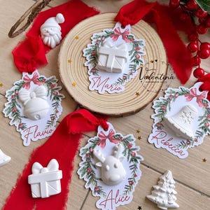 Könnte beinhalten: Weihnachtsanhänger mit weißen geprägten Designs eines Geschenks, einer Glocke, eines Rentiers, eines Weihnachtsmanns und eines Weihnachtsbaums. Jeder Anhänger hat ein rotes Band und ist mit einem Kranz und den Namen Mario, Flavio, Alice und Rebecca verziert.