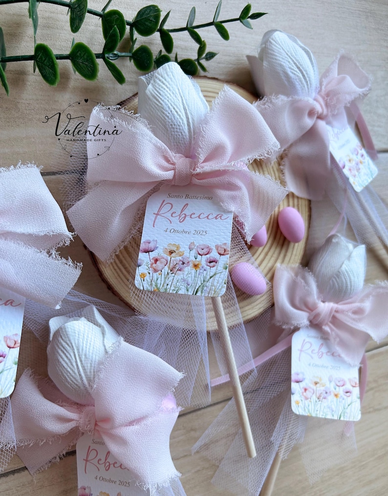 K&ouml;nnte beinhalten: Wei&szlig;e tulpenf&ouml;rmige Gastgeschenke mit rosa Schleifen und Etiketten mit der Aufschrift "Rebecca" und "4 Ottobre 2025". Die Gastgeschenke befinden sich auf Holzst&auml;ben und sind mit rosa T&uuml;ll verziert. Die Gastgeschenke befinden sich auf einer Holzscheibe.