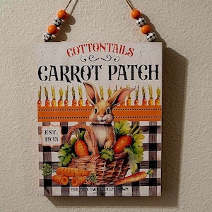 May include: A white wooden sign with the text "COTTONTAILS CARROT PATCH EST. 1933 PICK YOUR OWN LOCALLY GROWN" and a cartoon bunny rabbit holding a basket of carrots. The sign has a black and white checkered background.