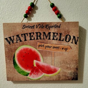 Pode incluir: Uma placa de madeira com fundo r&uacute;stico apresentando uma melancia com o texto "Sweet Vile Ripened Watermelon - pick your own - 4 mi" e um cord&atilde;o de contas vermelhas e verdes pendurado no topo.