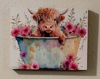 May include: A watercolor painting of a Highland cow taking a bath in a tub with pink flowers. The cow is brown and white with a flower crown on its head. The tub is blue and white with pink flowers around the rim.