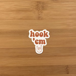May include: A sticker with the phrase "hook 'em" in orange, retro-style lettering. Below the text is a hand gesture with the index and pinky fingers extended. The sticker is on a wooden surface.