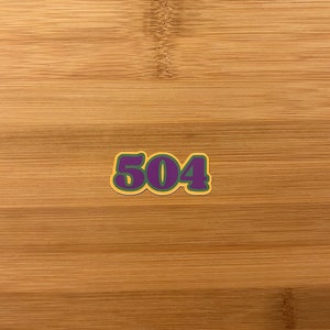 Puede incluir: Una pegatina con el número "504" en morado, contorneado en verde y con un borde amarillo. La pegatina está colocada sobre una superficie clara con vetas de madera.