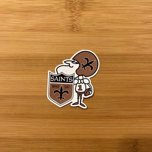 May include: A sticker featuring a cartoon football player in brown and white, wearing a helmet with a fleur-de-lis. The player holds a football and stands next to a shield with the text "SAINTS WHO DAT" and a fleur-de-lis symbol.