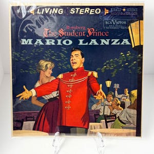 Pode incluir: Capa de álbum de vinil vintage para "The Student Prince" de Mario Lanza. A arte da capa apresenta um homem com um casaco vermelho de estilo militar com detalhes dourados, cantando. Uma mulher com uma saia vermelha e uma blusa branca está ao lado dele. O álbum é rotulado "Living Stereo."