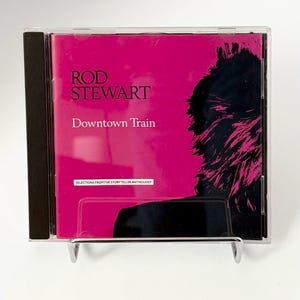 Puede incluir: Una caja de CD con una cubierta rosa brillante. El texto en la cubierta dice "ROD STEWART Downtown Train". El CD está en una caja de plástico transparente. La cubierta presenta una silueta negra del cabello y la cara de una persona.