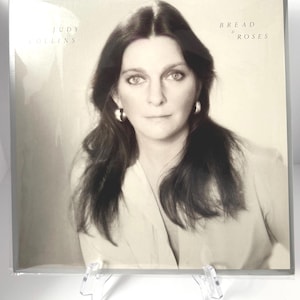 Puede incluir: Una portada de álbum en blanco y negro con Judy Collins. El título del álbum "Bread & Roses" está impreso en una fuente serif. La portada es un retrato de una mujer con pelo largo y oscuro y pendientes de aro. El álbum se muestra en un soporte de plástico transparente.