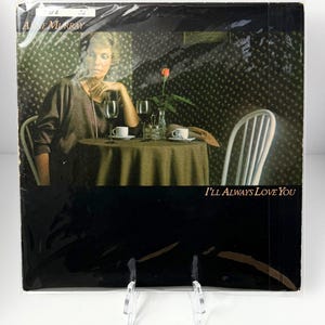 Puede incluir: Portada del álbum "I'll Always Love You" de Anne Murray. La imagen muestra a una mujer sentada a una mesa con una rosa, copas de vino y tazas de café. La portada está en una funda de plástico y se muestra en un soporte transparente.