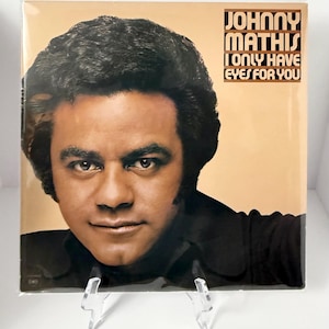 May include: Album cover of Johnny Mathis, with the text "Johnny Mathis I Only Have Eyes For You." The album art has a sepia tone with a close-up of Mathis's face. He is wearing a black shirt and has dark hair.