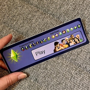 May include: A purple and blue sticker with a green diamond and the word "Play" in white. The sticker has a list of "Expansion Packs" and "Stuff Packs" with icons for each pack.