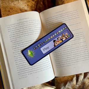 May include: A blue and green bookmark with the text "Expansion Packs: Play" and a photo of a group of people. The bookmark is lying on top of an open book with the text "The Bird King" on the page.