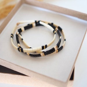 May include: Three beaded bracelets in a gift box. The bracelets feature a repeating pattern of black, cream, and gold-colored beads. The bracelets are resting in a light brown box with a white interior.