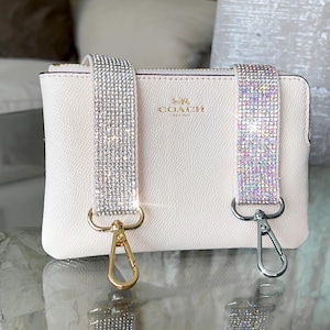 May include: A light pink Coach clutch with two detachable straps. One strap is silver with clear rhinestones, and the other is iridescent. The straps attach with gold and silver clasps.