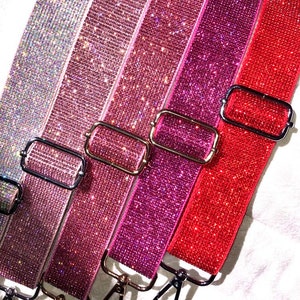 May include: Close-up of several sparkling, bedazzled bag straps in shades of pink, red, and silver. Each strap features a metal buckle and clasp. The straps are approximately 1.5 inches wide.