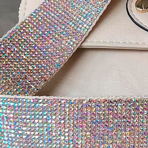 May include: Close-up of a cream-colored handbag with a sparkling, iridescent rhinestone-covered strap. The strap is wide and features a dense pattern of small, colorful rhinestones. A gold-colored clasp is visible on the bag.