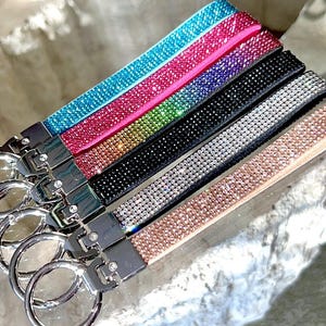 May include: A collection of keychains with wrist straps in various colors, including blue, pink, green, black, silver, and rose gold. Each strap is covered in sparkling rhinestones and attached to a silver-colored metal clasp with a key ring.