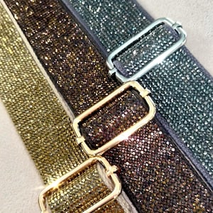 May include: Close-up of three shimmering straps with rectangular metal buckles. The straps are covered in small, sparkling embellishments in gold, brown, and silver. The buckles are gold and silver.