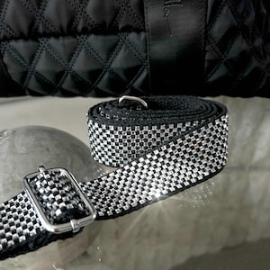 May include: Black and silver checkered strap with a silver buckle. The strap is rolled up and also attached to a black quilted bag. The strap is made of a black material with silver square accents.