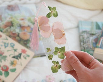 May include: A clear, rectangular bookmark with pressed pink flower petals and green leaves. A pink tassel hangs from the top. Other floral designs are visible in the background.