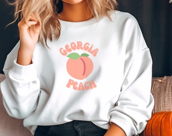 Könnte beinhalten: Weißer Pullover mit einem Pfirsich-Motiv und dem Text "Georgia Peach" in einer Retro-Schrift.
