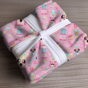 May include: A pink robe with a repeating pattern of nutcrackers, drums, candy canes, and snowflakes. The robe is tied with a white belt. The design is festive and holiday-themed.