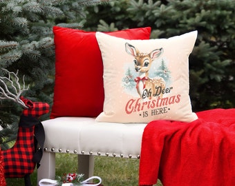 May include: A cream-colored throw pillow with a vintage illustration of a deer with a red bow. The text "Oh Deer Christmas is Here" is printed in red on the pillow.