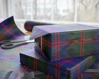 Puede incluir: Dos regalos envueltos en papel a cuadros azul, verde y rojo están apilados sobre una mesa. Se ven un rollo de papel a juego y unas tijeras. Una cinta gris está en primer plano. La escena está ambientada cerca de una ventana.