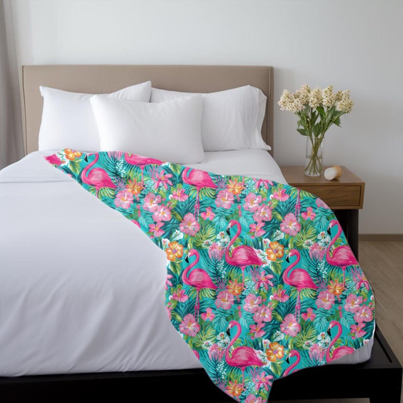 May include: A turquoise blue throw blanket with a pink flamingo and tropical floral pattern. The blanket is folded on a white bed with a pink headboard.