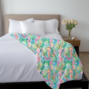 Op de afbeelding: Een wit bed met een lichtblauw dekbedovertrek met een herhalend patroon van roze, gele en blauwe zeepaardjes en groene zeewier.