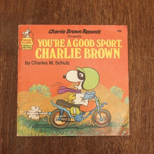 May include: A red and orange record cover featuring Snoopy riding a motorcycle. The cover has the text "Charlie Brown Records Presents You're a Good Sport, Charlie Brown" and "by Charles M. Schulz".