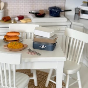 May include: Miniature kitchen scene with white cabinets, a table, and chairs. A stack of food containers sits on the table, along with a plate of pasta and bread. A small rug is under the table. The kitchen has a stove, sink, and other accessories.