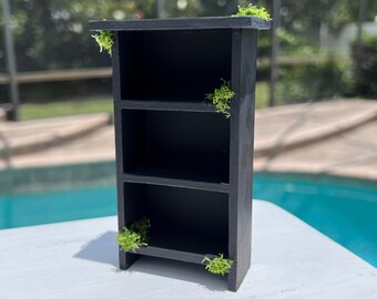 May include: A black wooden bookshelf with three shelves. The bookshelf has green moss accents on the top and sides.