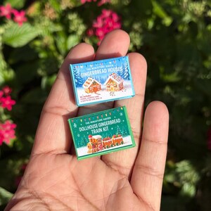 May include: Two miniature kits for gingerbread houses and a train, held in a hand. The gingerbread house kit is in a blue box with a winter scene. The train kit is in a green box with a train illustration. Both kits are labeled "The Miniature Haven".
