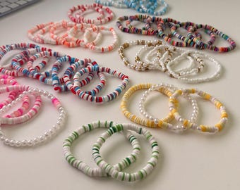 Op de afbeelding: Een verzameling kleurrijke armbanden in verschillende ontwerpen. De armbanden hebben een mix van witte, roze, rode, blauwe, groene en gele kralen, sommige met parelaccenten. De armbanden zijn gerangschikt op een wit oppervlak.