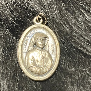 May include: A silver metal pendant with a raised image of Saint Faustina Kowalska. The pendant is oval shaped and has a loop at the top for hanging.