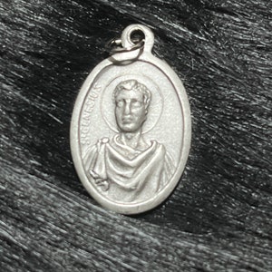 May include: Oval silver pendant with a portrait of a man with a halo, and the inscription "GENESIUS". The pendant has a small loop at the top for attaching to a chain. The pendant is a religious item.