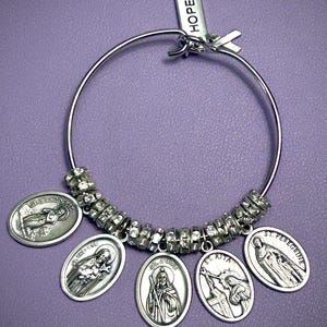 May include: Silver-toned bangle bracelet with religious medallions. The bracelet features oval charms with images of saints, including St. Jude and St. Rita. A small tag with the word "HOPE" is attached. The bracelet is set against a purple background.