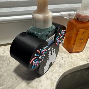 May include: Black soap dispenser holder with a Disney castle and fireworks design. The holder is designed to hold a soap dispenser. The holder has a black base with a white castle and pink and blue fireworks.