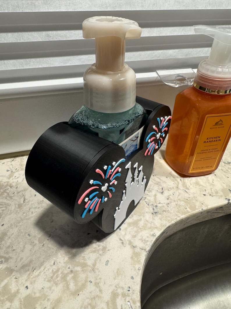 May include: Black soap dispenser holder with a Disney castle and fireworks design. The holder is designed to hold a soap dispenser. The holder has a black base with a white castle and pink and blue fireworks.