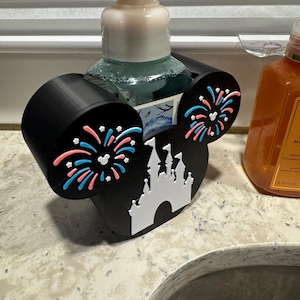 May include: Black soap dispenser holder shaped like Mickey Mouse ears, with a white castle and fireworks design. The holder is designed to hold a soap dispenser bottle. The ears have pink and blue fireworks.