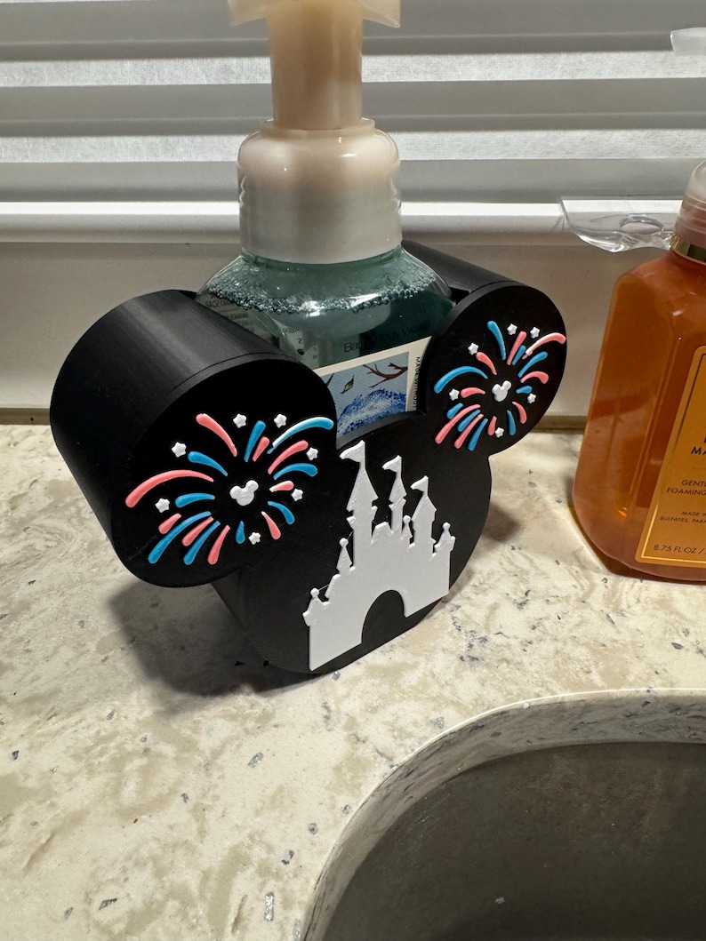 May include: Black soap dispenser holder shaped like Mickey Mouse ears, with a white castle and fireworks design. The holder is designed to hold a soap dispenser bottle. The ears have pink and blue fireworks.