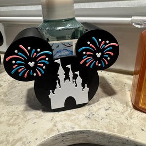 May include: Black Mickey Mouse-shaped soap dispenser holder with white castle and fireworks design. The ears feature pink and blue fireworks. A decorative item for the bathroom.