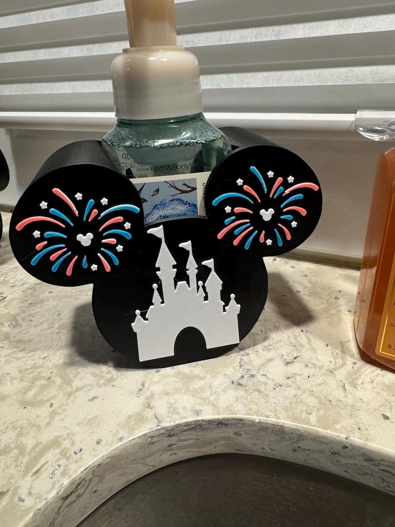 May include: Black Mickey Mouse-shaped soap dispenser holder with white castle and fireworks design. The ears feature pink and blue fireworks. A decorative item for the bathroom.