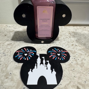 May include: A black Mickey Mouse-shaped hand soap holder with a pink bottle of "Champagne Toast" foaming hand soap. The holder is on a countertop with a matching coaster featuring a white castle and fireworks.