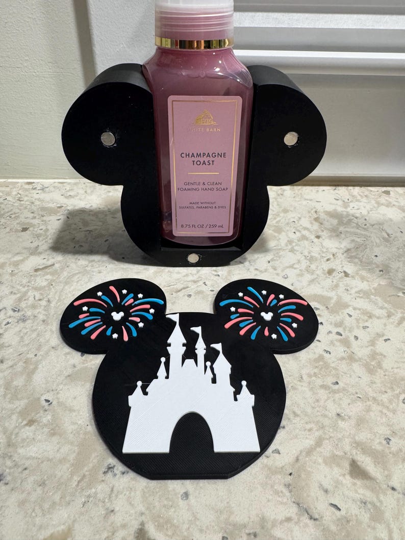 May include: A black Mickey Mouse-shaped hand soap holder with a pink bottle of "Champagne Toast" foaming hand soap. The holder is on a countertop with a matching coaster featuring a white castle and fireworks.