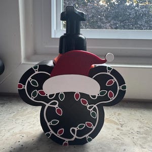 May include: A black soap dispenser shaped like a mouse head, featuring a red and white Santa hat on top. The black surface is decorated with a white string of Christmas lights with small red and green bulbs. A black pump mechanism is attached to the top.