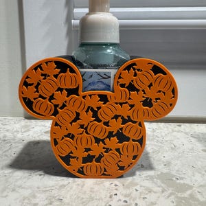 May include: A soap dispenser holder shaped like a mouse head silhouette, featuring a black base with an orange overlay of pumpkins and maple leaves. The holder contains a clear plastic bottle with a white pump top and a partial label.